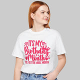 A woman with red hair wearing a white graphic t-shirt and light blue jeans, standing against a plain white background. The t-shirt features a pink graphic that reads 'IT'S MY Birthday Months BE I GET THE WHOLE MONTH'.