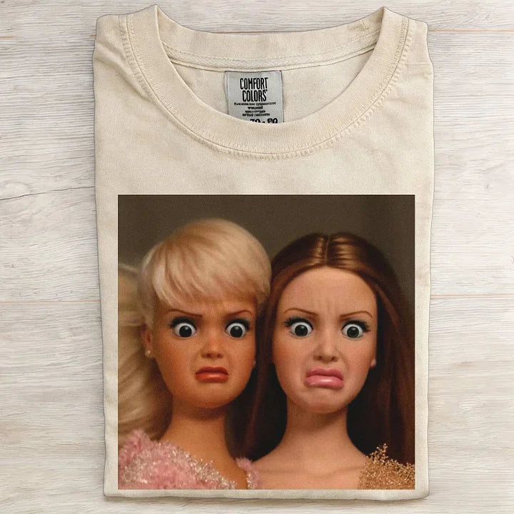 Beige t-shirt with a graphic of two dolls with exaggerated facial expressions on a wooden surface.